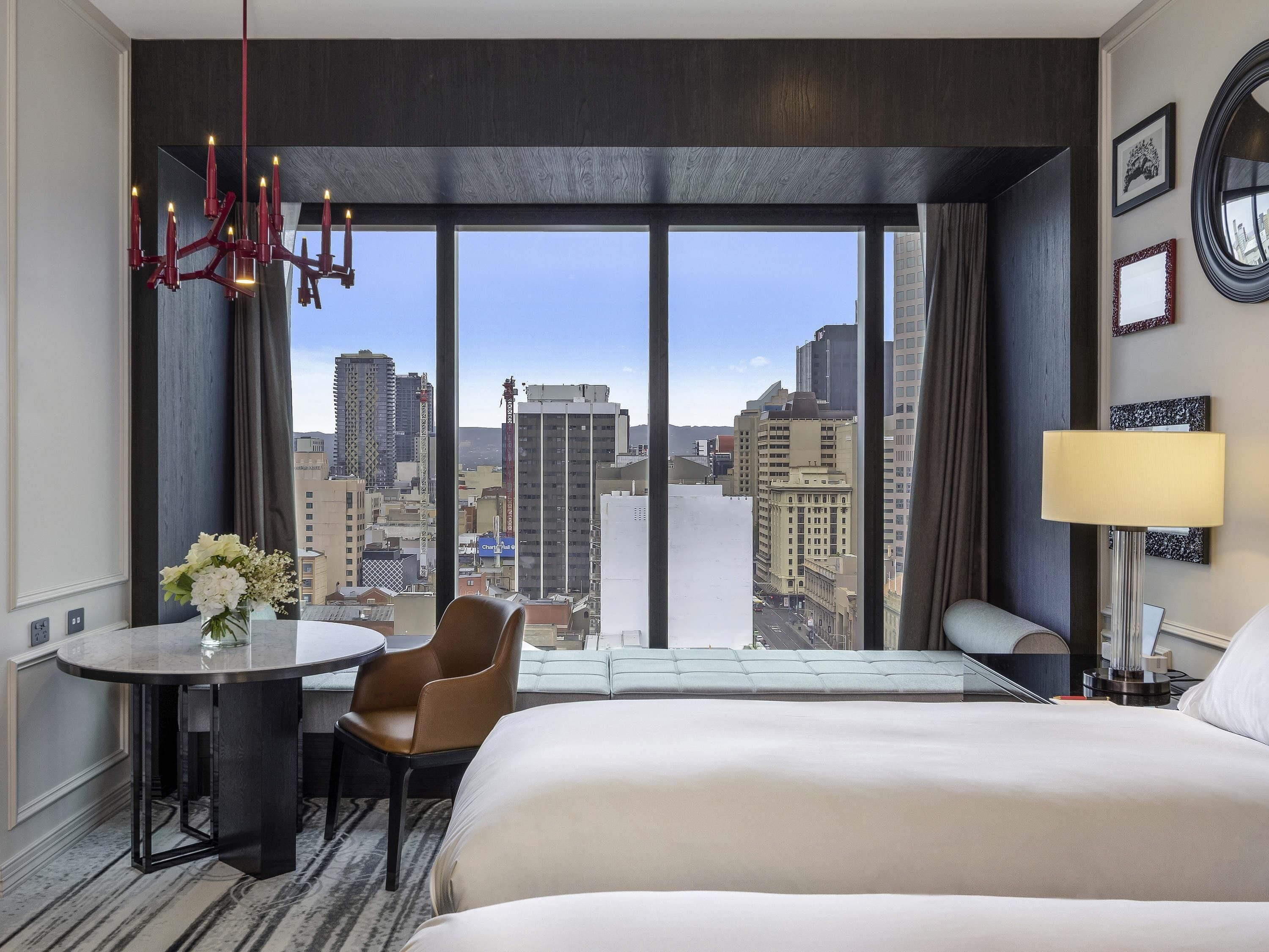 A hotel room with a large bed, a desk with a chair, a round table with a vase of flowers, a lamp, and a view of the cityscape through large windows.