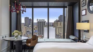 A hotel room with a large bed, a desk with a chair, a round table with a vase of flowers, a lamp, and a view of the cityscape through large windows.