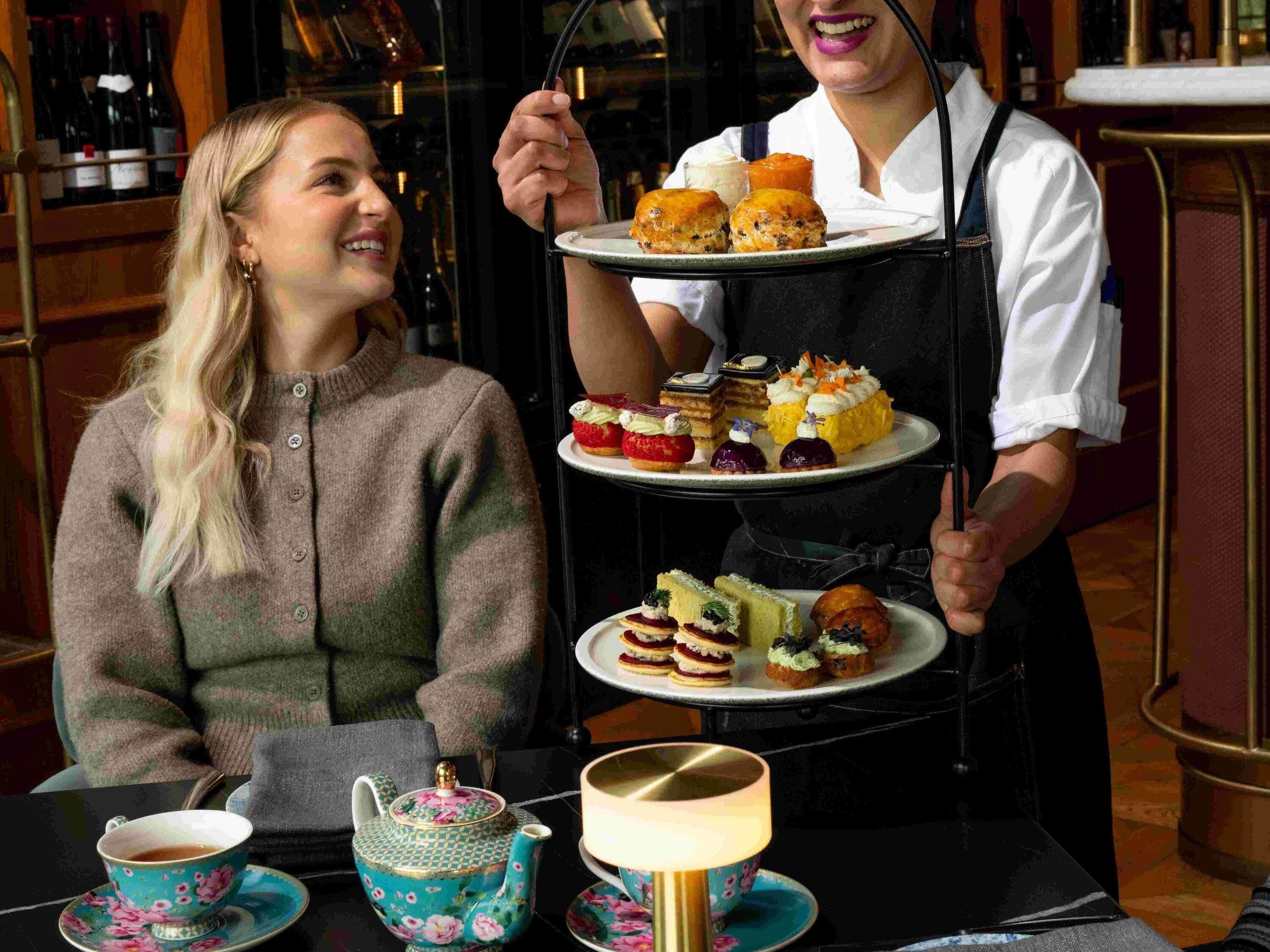 Une personne tenant un plateau à étages sur lequel sont disposés divers gâteaux et desserts, une théière et une tasse, le tout posé sur une table.