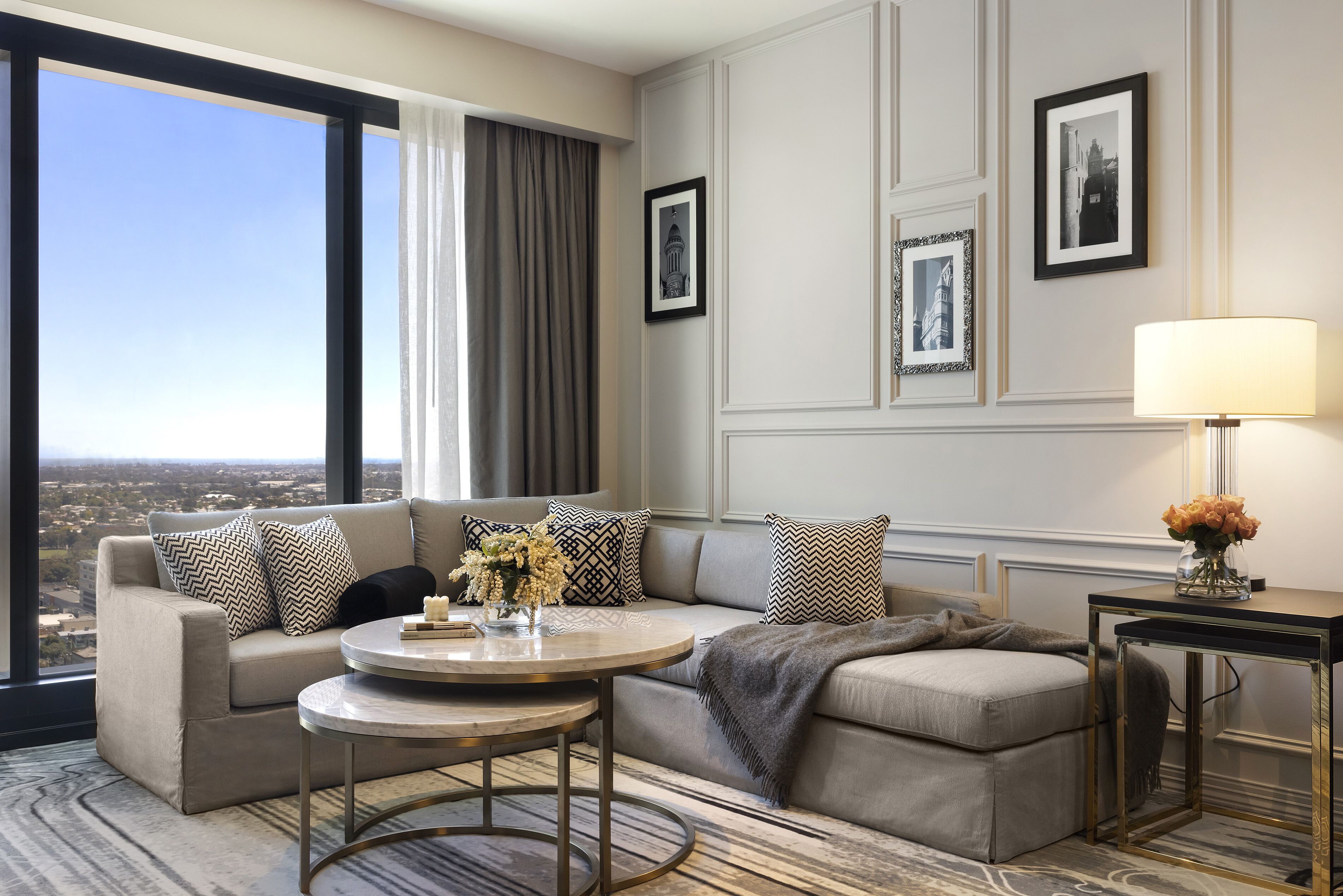 A modern hotel room with a large window, a sofa set, a round coffee table, and a side table with a lamp.