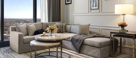 A modern hotel room with a large window, a sofa set, a round coffee table, and a side table with a lamp.