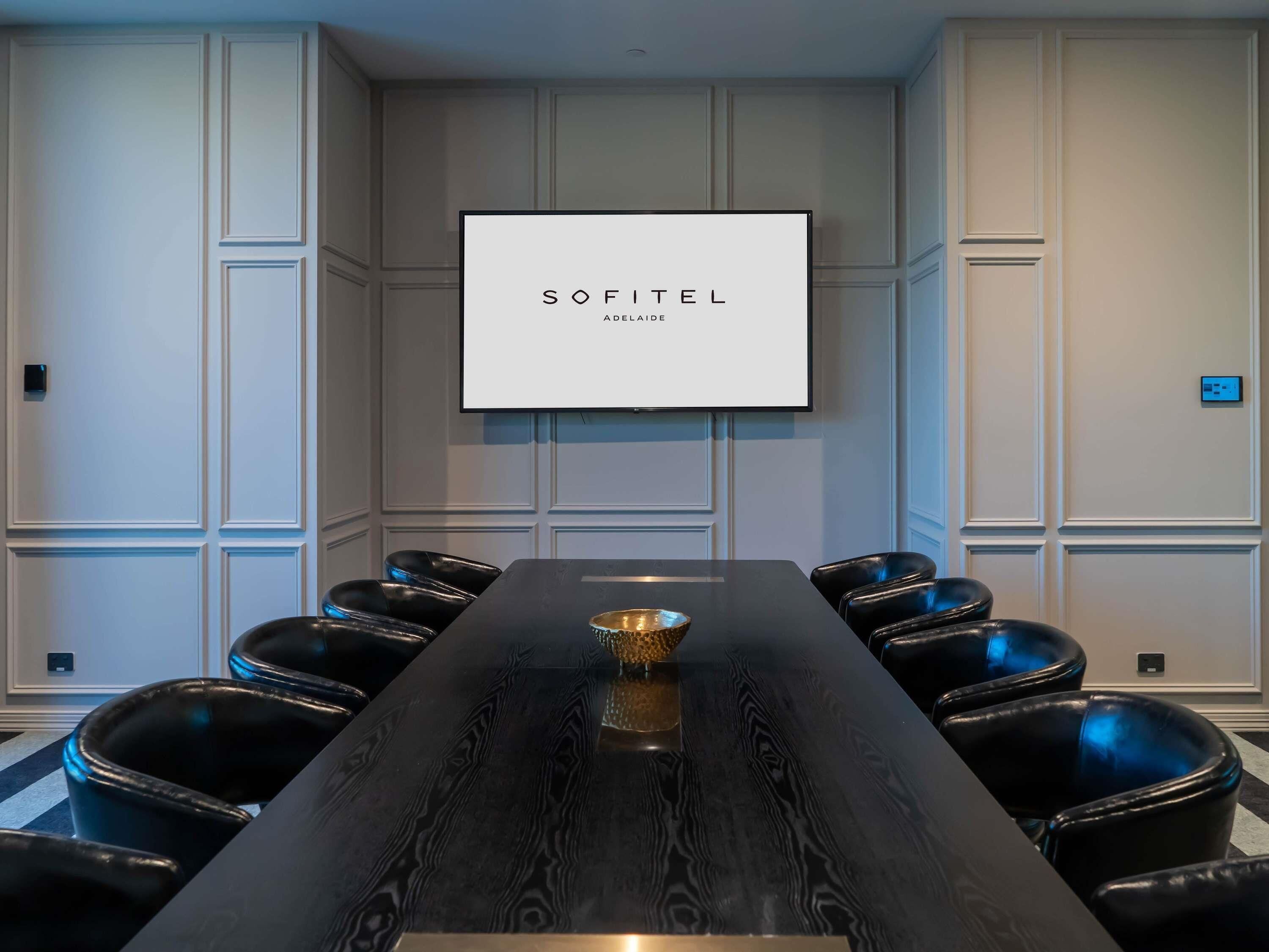 A conference room with a long table, black leather chairs, a screen displaying 'SOFITEL ABELATIDE,' and white paneled walls.