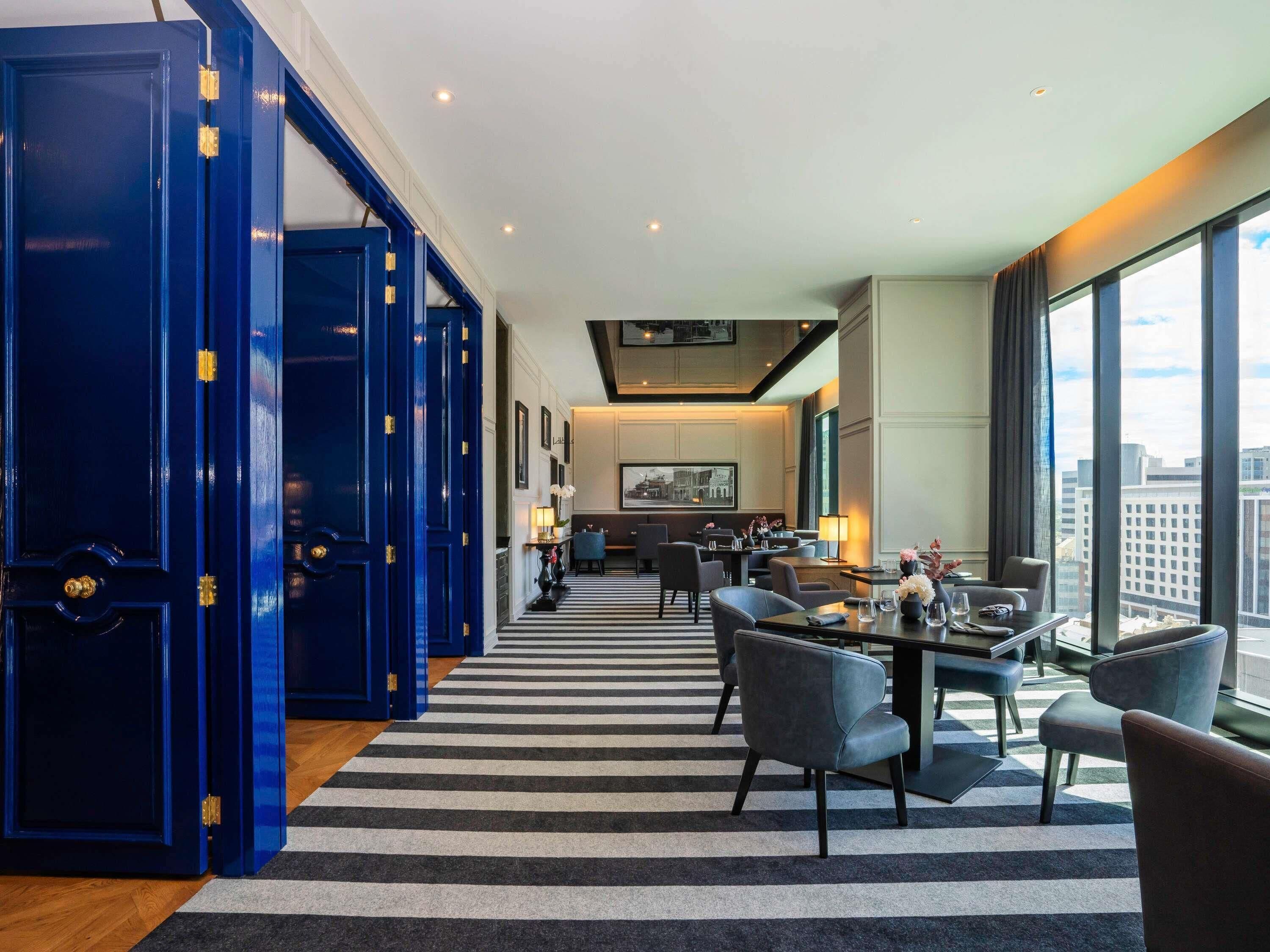 A hotel lobby with a striped carpet, blue doors, a dining area with tables and chairs, and a city view through large windows.