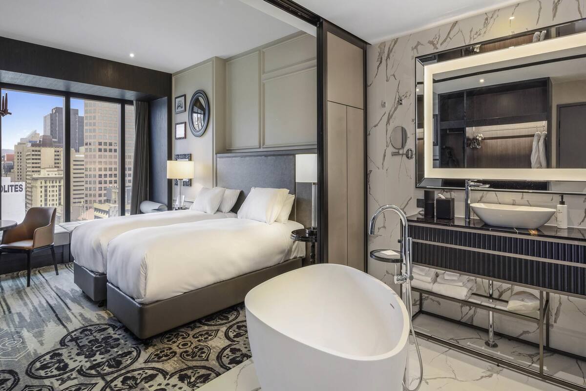 A modern hotel room with a large bed, a bathtub, a vanity with a large mirror, and a city view.