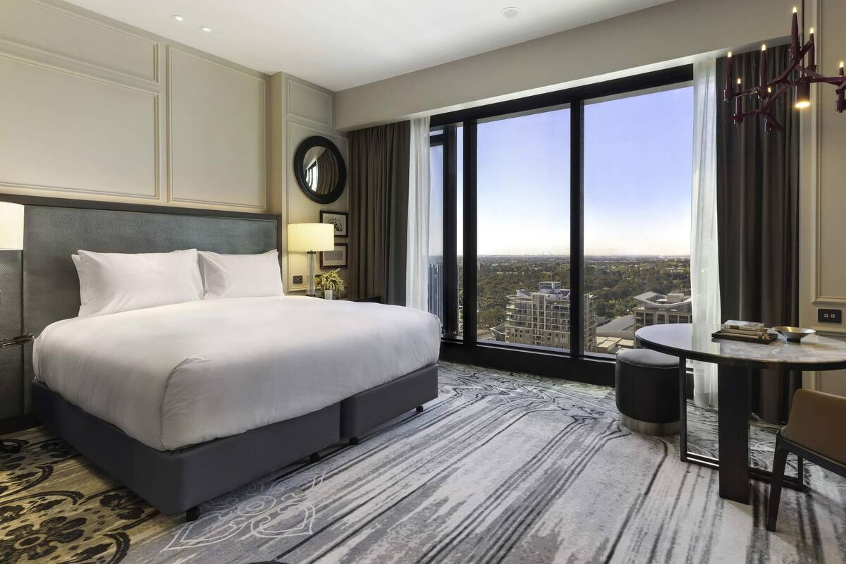 A hotel room with a large bed, a round mirror, a desk with a chair, and a view of the city through large windows.