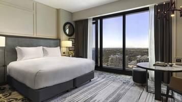 A hotel room with a large bed, a round mirror, a desk with a chair, and a view of the city through large windows.