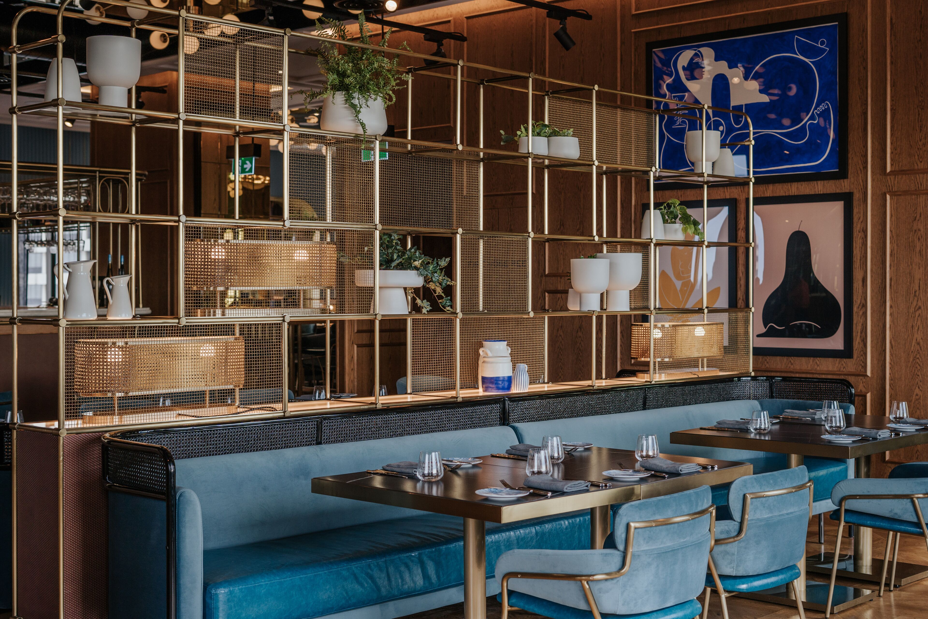 L’intérieur d’un restaurant présente des sièges bleus rembourrés, des étagères en métal ornées de plantes et des œuvres d’art accrochées aux murs.