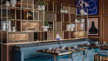 Ein Restaurantinnenraum mit blau gepolsterten Sitzmöglichkeiten, Metallregalen mit Pflanzen und Kunstwerken an den Wänden.