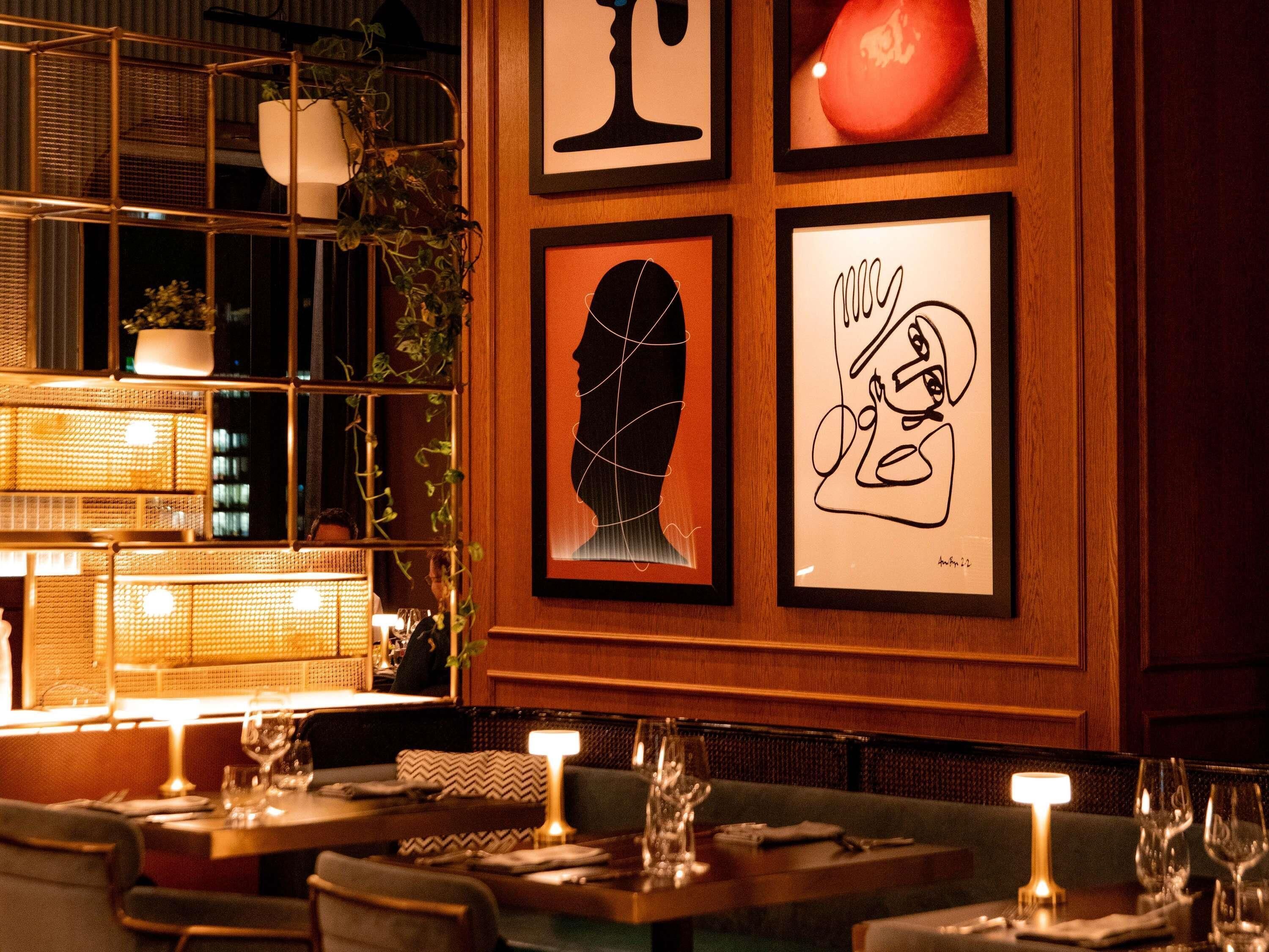 Ein Restaurantinnenraum mit gerahmten Kunstwerken an der Wand, gedeckten Tischen mit Gläsern und Besteck, und warmem, indirektem Licht.
