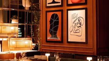 Ein Restaurantinnenraum mit gerahmten Kunstwerken an der Wand, gedeckten Tischen mit Gläsern und Besteck, und warmem, indirektem Licht.