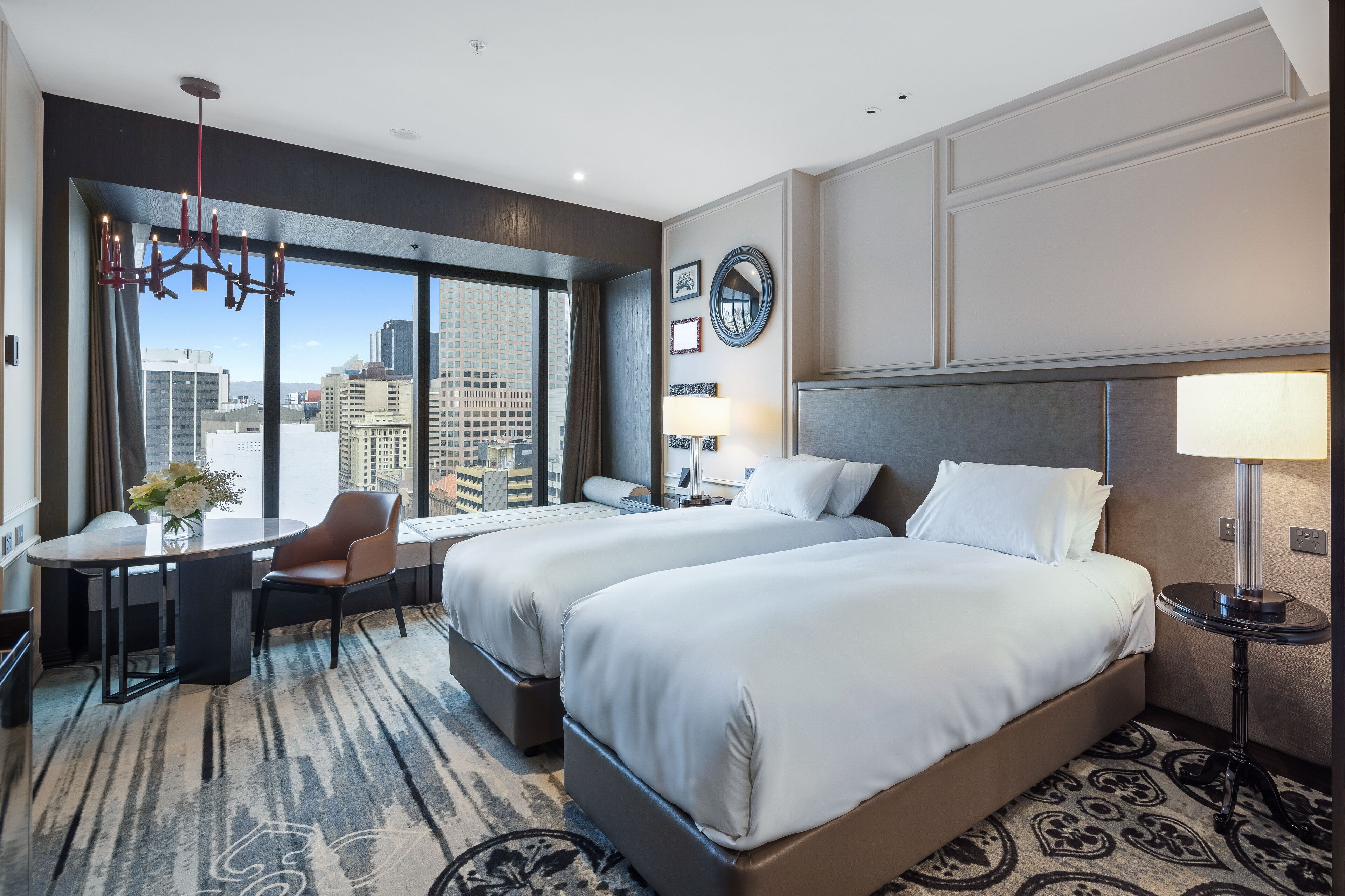 Une chambre d’hôtel avec deux lits, un bureau, une chaise, un lustre, un miroir rond et une vue sur la ville.