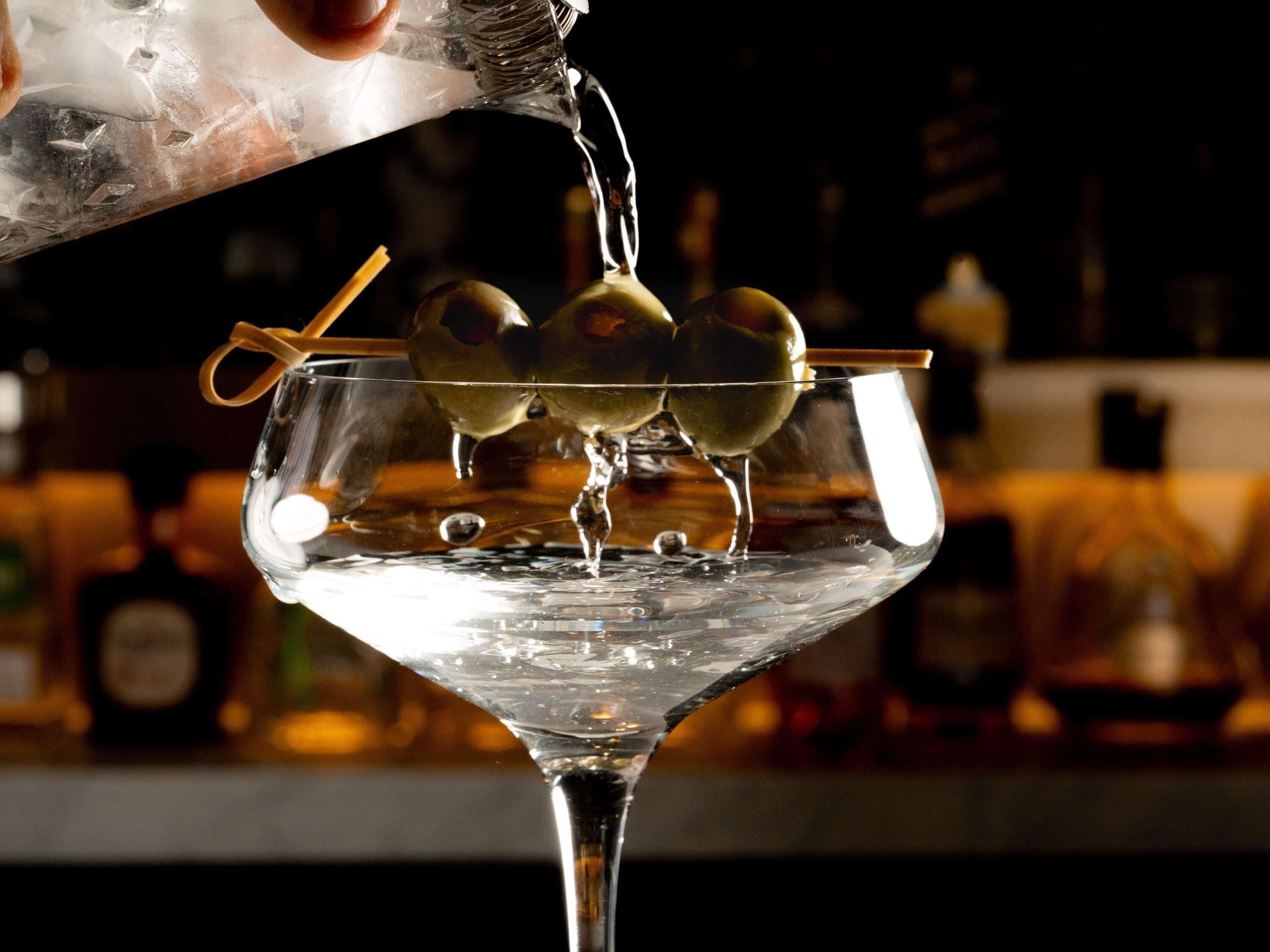 A martini glass with olives and a straw, with a hand pouring liquid into it.