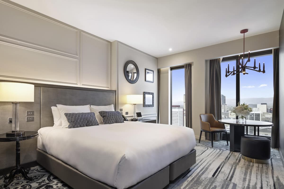 A hotel room with a large bed, a desk, a chair, a view of the city, and a chandelier.