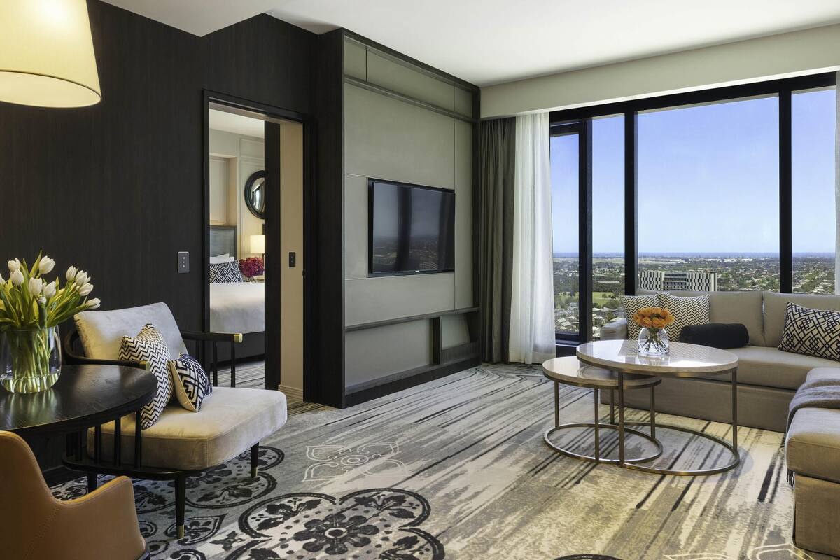 A modern hotel room with a large window, a flat-screen TV, a sofa, a coffee table, and a decorative rug.