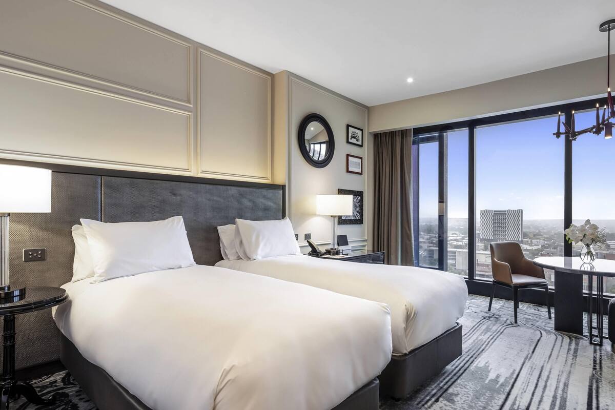 A hotel room with two beds, a desk, a chair, a large window with a city view, and a chandelier.