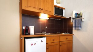 A compact kitchen with wooden cabinets, a microwave, a refrigerator, and a small shelf with kitchen items.