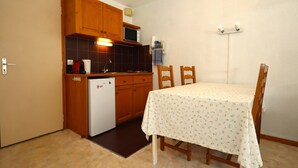 A compact kitchen with wooden cabinets, a refrigerator, and a microwave. There is a dining table with chairs and a small rug.