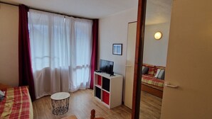 Ett litet, väl upplyst rum med en säng, en tv på ett stativ, ett litet bord och en stol.