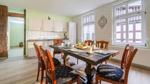 A dining area with a wooden table and chairs, a kitchen in the background, and large windows with shutters. - Ferienwohnung Fachwerktraum, Wernigerode (Wernigerode)