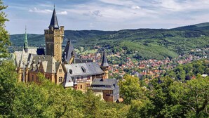 A historic building with a prominent tower and multiple spires, surrounded by lush green hills and a small town below. - Ferienwohnung Fachwerktraum, Wernigerode (Wernigerode)