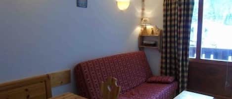 Um quarto aconchegante com uma mesa de madeira, um sofá estampado, um pequeno aparador com objetos decorativos e uma janela com cortinas.