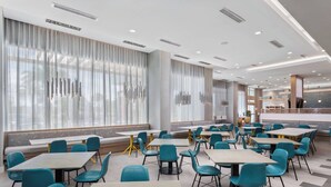 Un comedor moderno con varias mesas y sillas, ventanas amplias con cortinas, y una zona de estar con bancos.