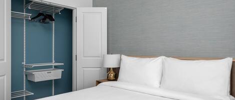 Een opgemaakt bed met witte lakens, een bedlampje en een wandplank met hangende kleding.
