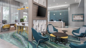 Un lobby moderno de hotel con un mostrador de recepción, un área de descanso con sillas y mesas, una gran pantalla de televisión y un letrero que dice 'Hello Orlando'.