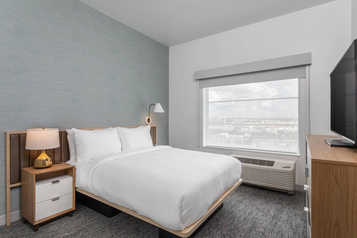 Habitación de hotel con cama, mesita de noche con lámpara, televisor y ventana con vista a la ciudad.