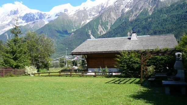 A rustic cabin with a sloped roof, surrounded by a wooden fence, set against a backdrop of snow-capped mountains and lush greenery.