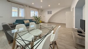 A modern living room with a glass dining table, white chairs, a blue sofa, and a TV on a wooden stand.