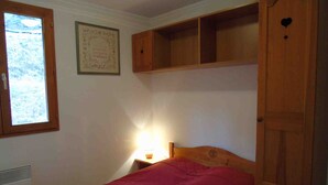 A bedroom with a single bed, a window, a wall-mounted shelf, and a bedside table with a lamp.