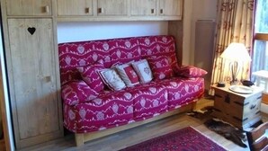 A cozy room with a wooden cabinet with heart-shaped cutouts, a patterned sofa, a small side table with a lamp, and a window with curtains. - Charming studio for 4 near slopes with balcony, pets allowed (Valfréjus)