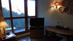 A cozy room with a large window, a television, a table, and a lamp. - Charming studio for 4 near slopes with balcony, pets allowed (Valfréjus)