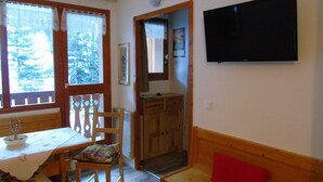 A cozy room with a wooden dining table, chairs, a bench, a TV mounted on the wall, and a door leading to another room. - Cozy studio 4 ppl, near gondola & shops (Valfréjus)