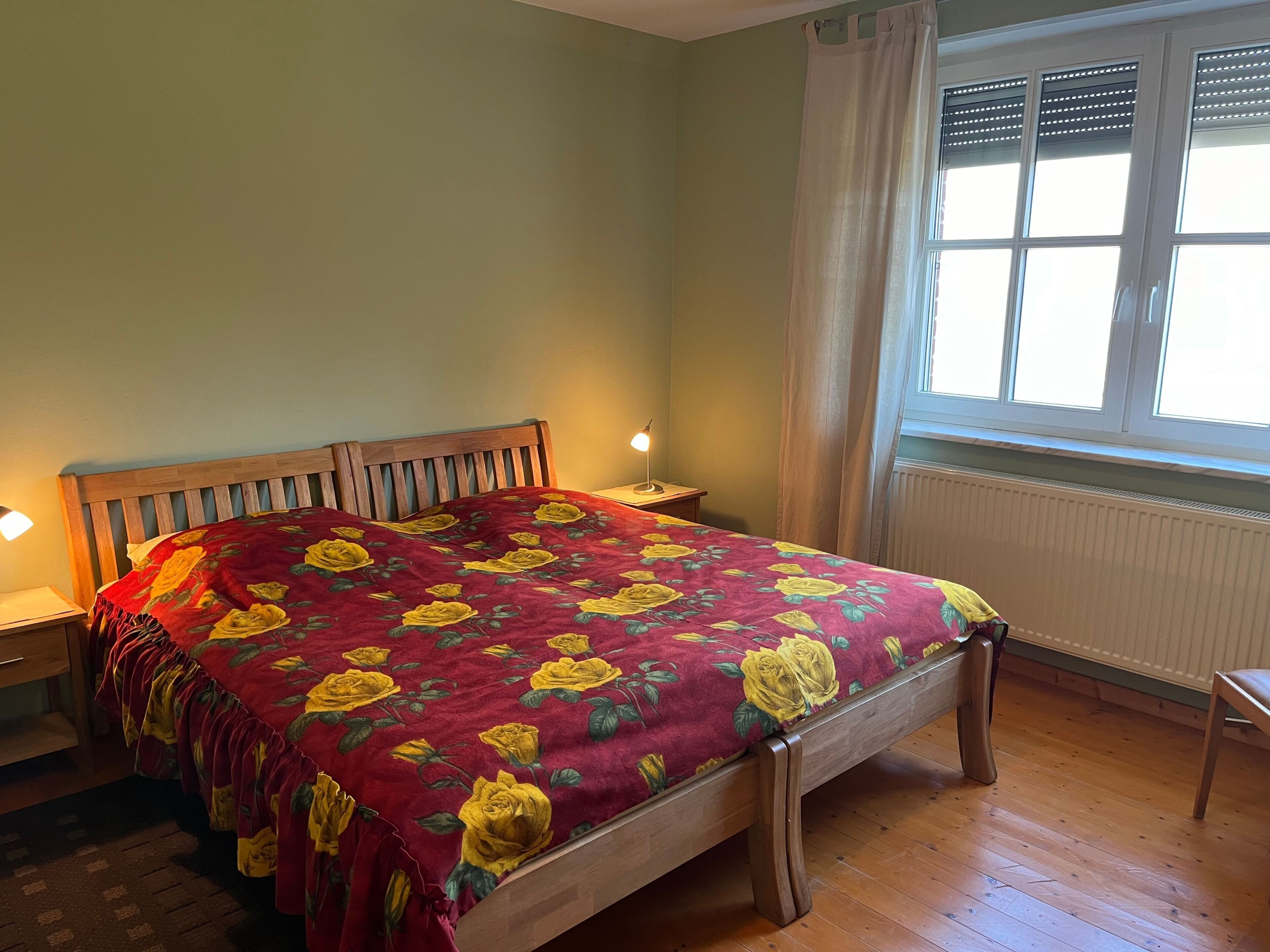 Une chambre avec un lit, une table de chevet, une lampe, une fenêtre avec des stores et un radiateur.