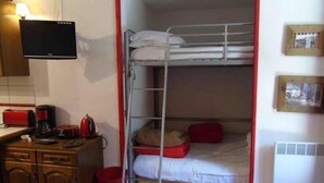 Une petite pièce compacte avec des lits superposés, un bureau sur lequel se trouve une bouilloire rouge, une télévision et une petite poubelle.