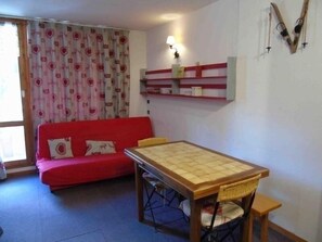A small living area with a red sofa, a wooden table, and a chair. There is a window with curtains, a wall-mounted shelf with items, and a wall-mounted lamp.