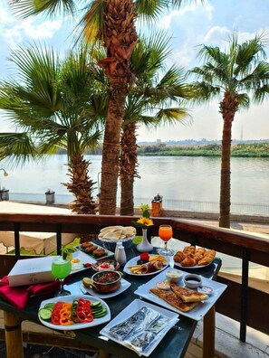 A table set with a variety of foods and drinks, overlooking a body of water with palm trees in the background. - Jewel Maadi Cabins and Club (Cairo)