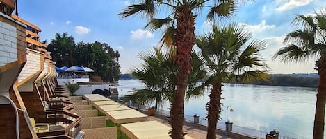 A terrace with outdoor seating, palm trees, and a view of a body of water.