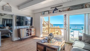 A cozy room with a wooden floor, a large TV, a wooden cabinet, and a view of the beach through a glass door. - Vintage Vacation Pet Friendly, Ocean Views! (Surf City)