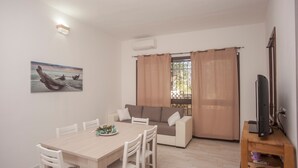 Una stanza con un tavolo da pranzo, sedie bianche, un divano, una TV su un mobile in legno e un quadro appeso al muro.
