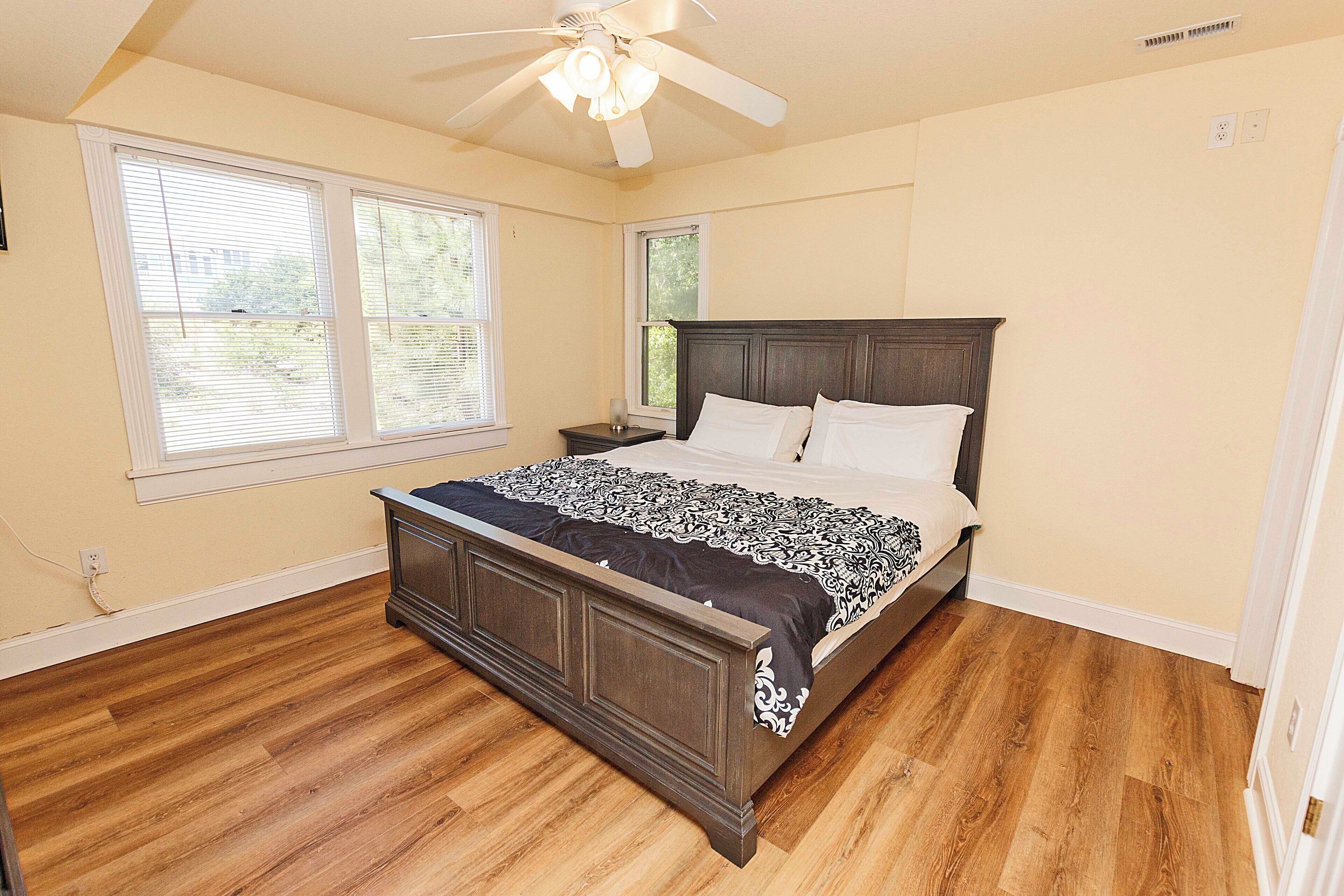 A bedroom with a wooden bed, two windows, a ceiling fan, and a nightstand.