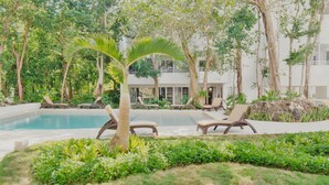 A pool area with lounge chairs, palm trees, and a building in the background. - Private Tropical Home  with Pool (Akumal)