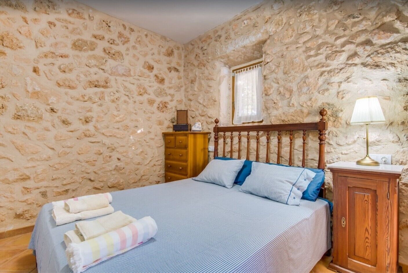 A bedroom with a stone wall, a wooden bed frame, a bedside table, and a lamp.