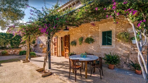 A rustic stone building with a round table and chairs outside, surrounded by potted plants and flowering vines. - Beautiful Country Home “Son Vanrell” with Mountain View, Pool, Wi-Fi, A/C, Terrace & Garden; (Campos)