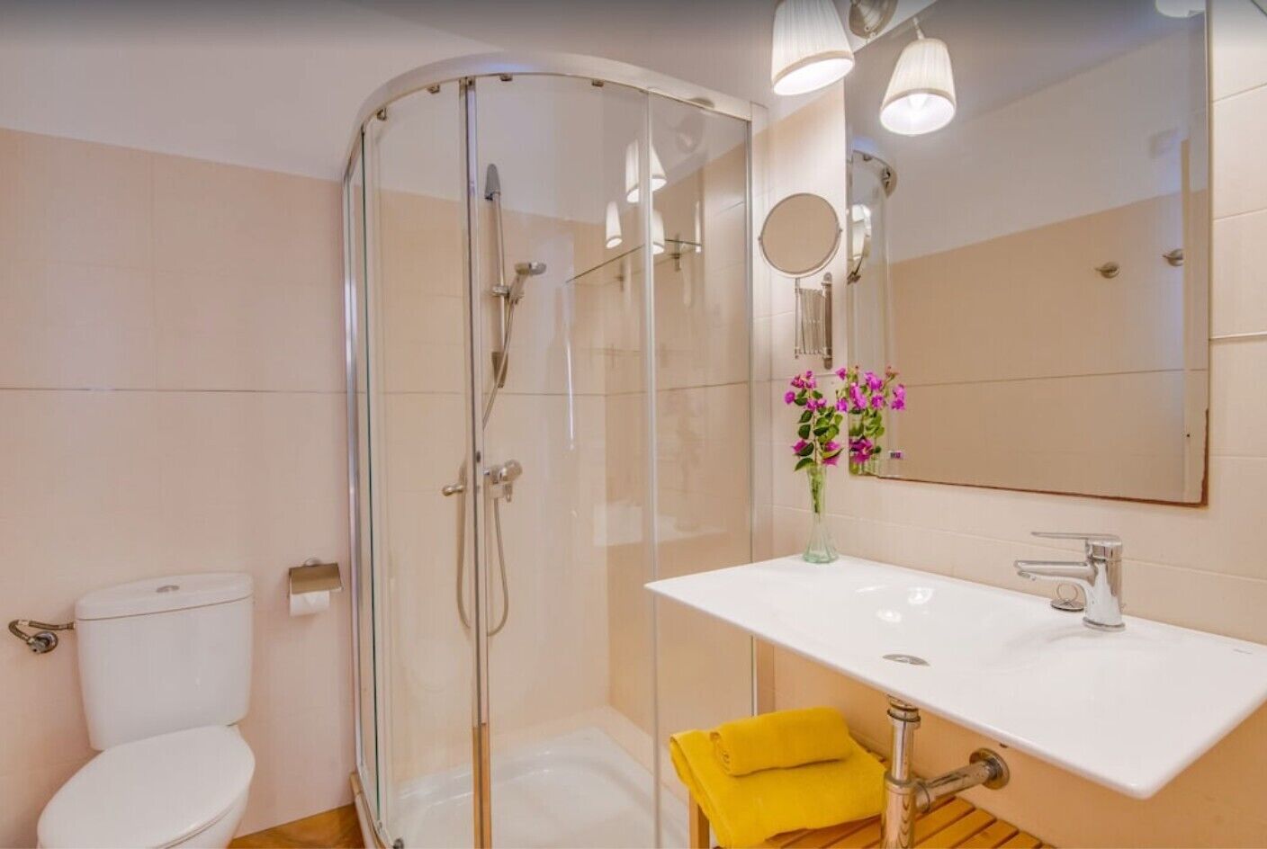 A bathroom with a glass-enclosed shower, a toilet, a sink with a mirror, and a vase with flowers.