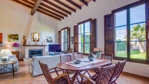 A spacious living area with a fireplace, television, dining table, and large windows offering a view of the outdoors. - Beautiful Country Home “Son Vanrell” with Mountain View, Pool, Wi-Fi, A/C, Terrace & Garden; (Campos)