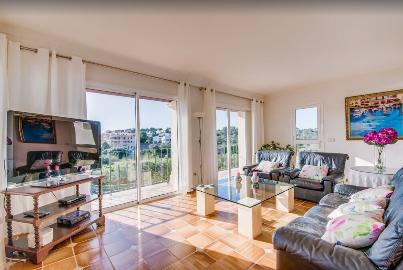 A spacious living room with a large TV, leather sofas, a glass coffee table, and a balcony with a scenic view.