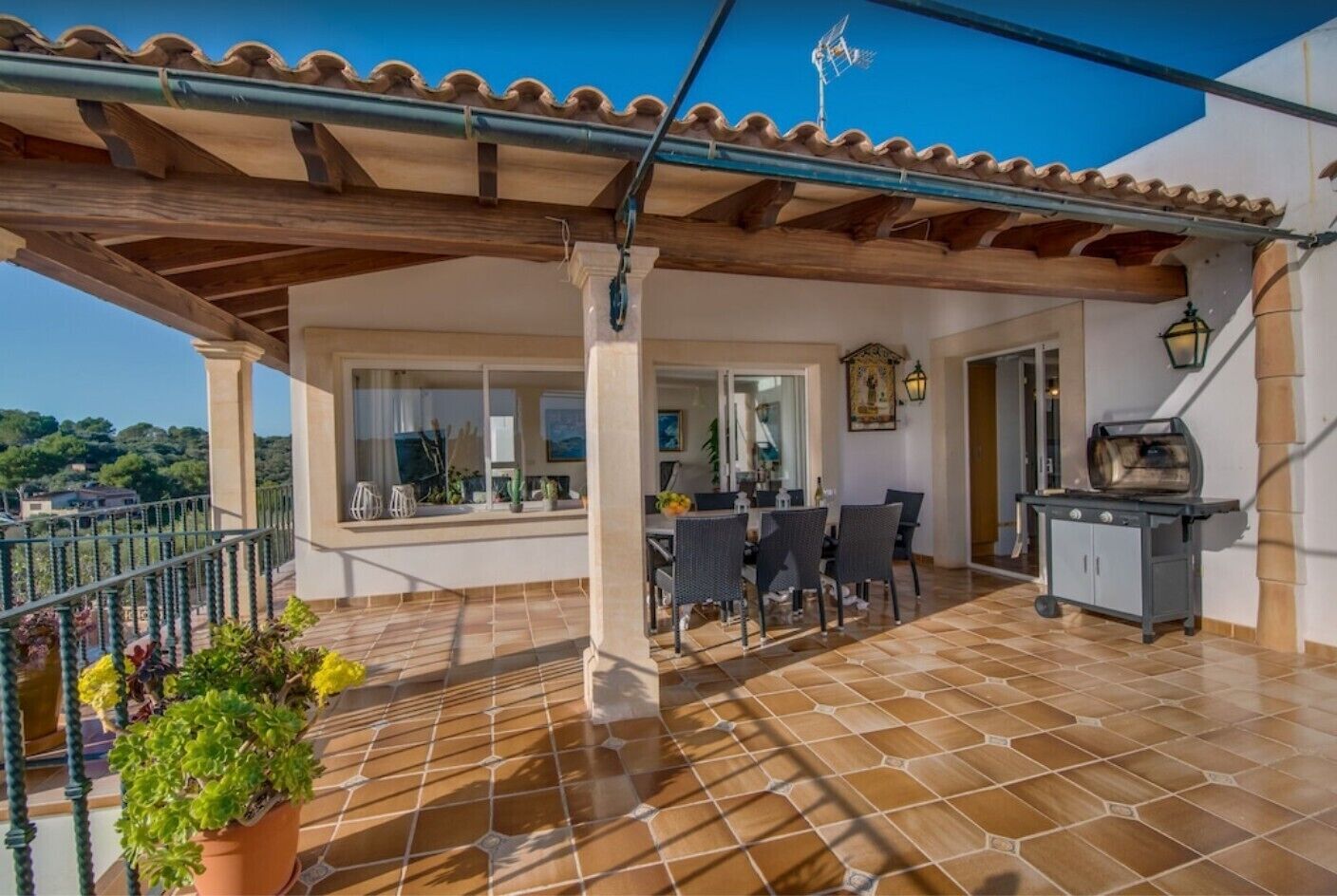 Un patio techado con suelo de baldosas, una mesa de comedor con sillas, una parrilla y plantas en macetas.